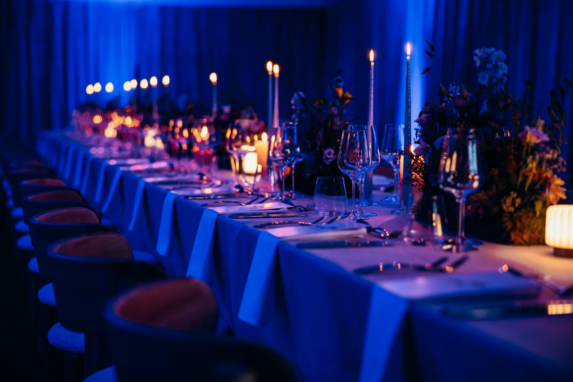 Long banquet table set with candles, glassware and florals under deep blue lighting at an Archer evening event