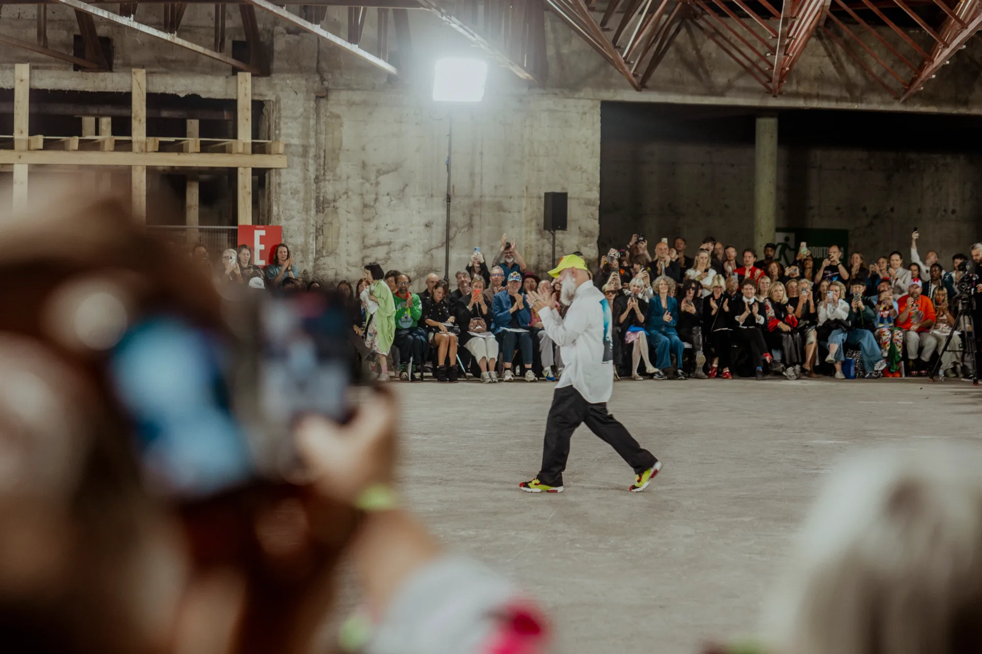 Designer walking the runway of a converted warehouse while the seated audience applauds at Antwerp Fashion Show