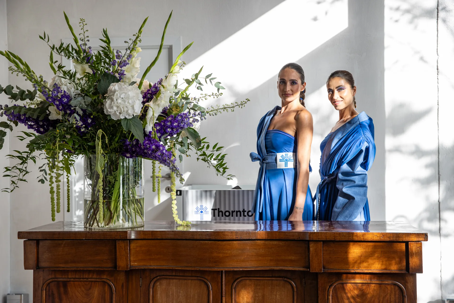 Two models in blue Thornton outfits beside a large floral arrangement at a Thornton brand event