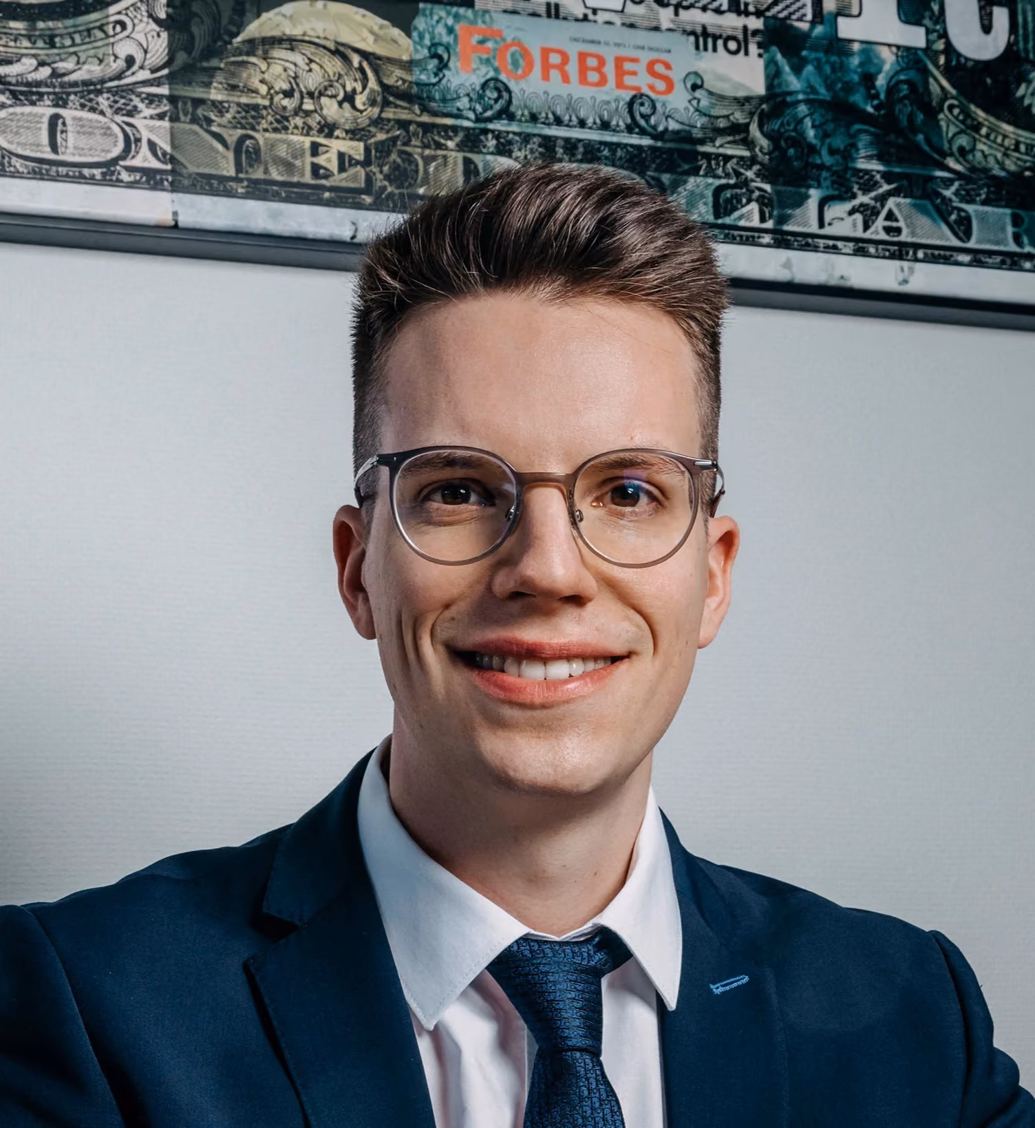 Smiling man with glasses wearing a navy suit and tie in front of a wall with Forbes decor.