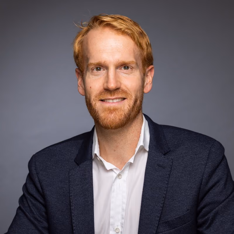 Smiling red-haired man with beard wearing white shirt and dark blazer on gray background.