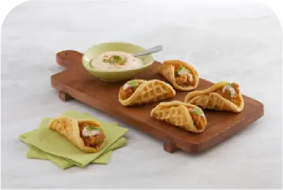 Mini waffle tacos filled with chicken and garnished with sauce and herbs on a wooden board with a dipping bowl.