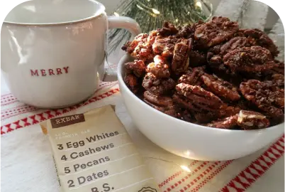 White bowl of candied pecans, a recipe card, and a white cup with 'MERRY' on a festive cloth.