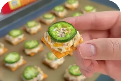 Hand holding a bite-sized cracker topped with cheese, tuna salad, and a jalapeño slice.