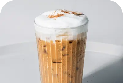 Tall glass of iced coffee topped with thick foam and a dusting of cinnamon or cocoa powder.