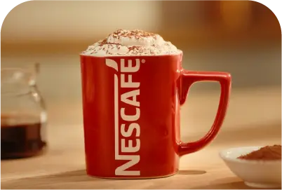 Red NESCAFÉ mug filled with whipped cream topped coffee on a light wooden table.