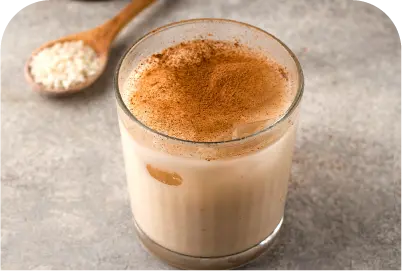Glass of horchata topped with cinnamon powder on a gray surface with a wooden spoon of rice.