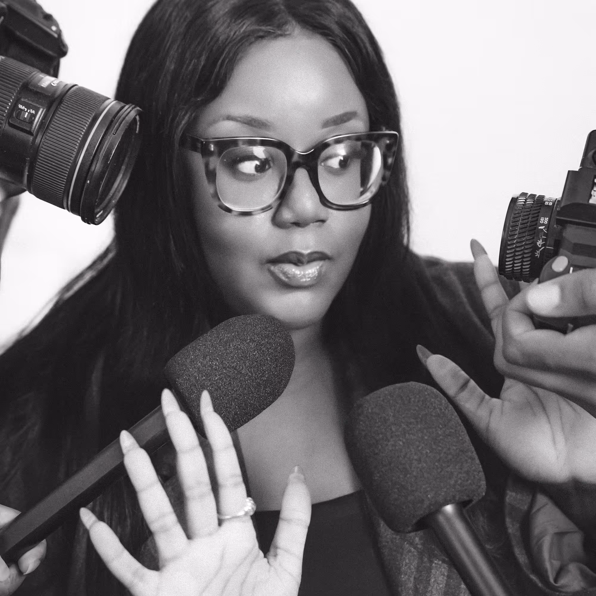 Woman with glasses facing multiple microphones and cameras, gesturing with her hands.
