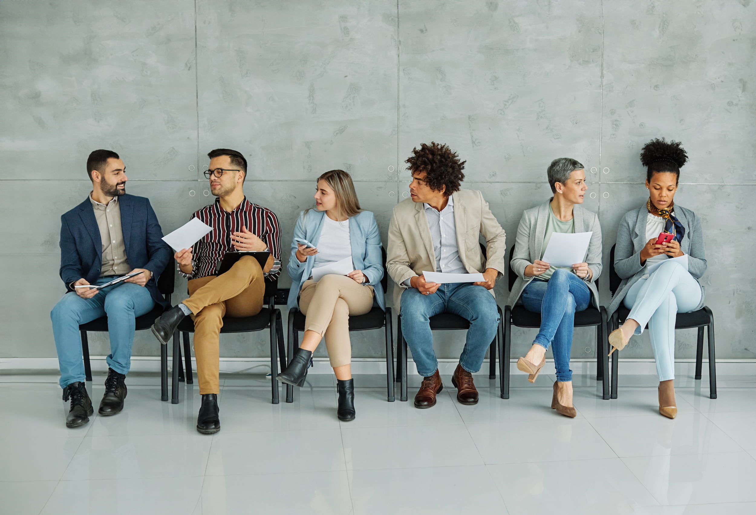 Six diverse adults seated in a row, holding papers, waiting against a gray wall on black chairs.