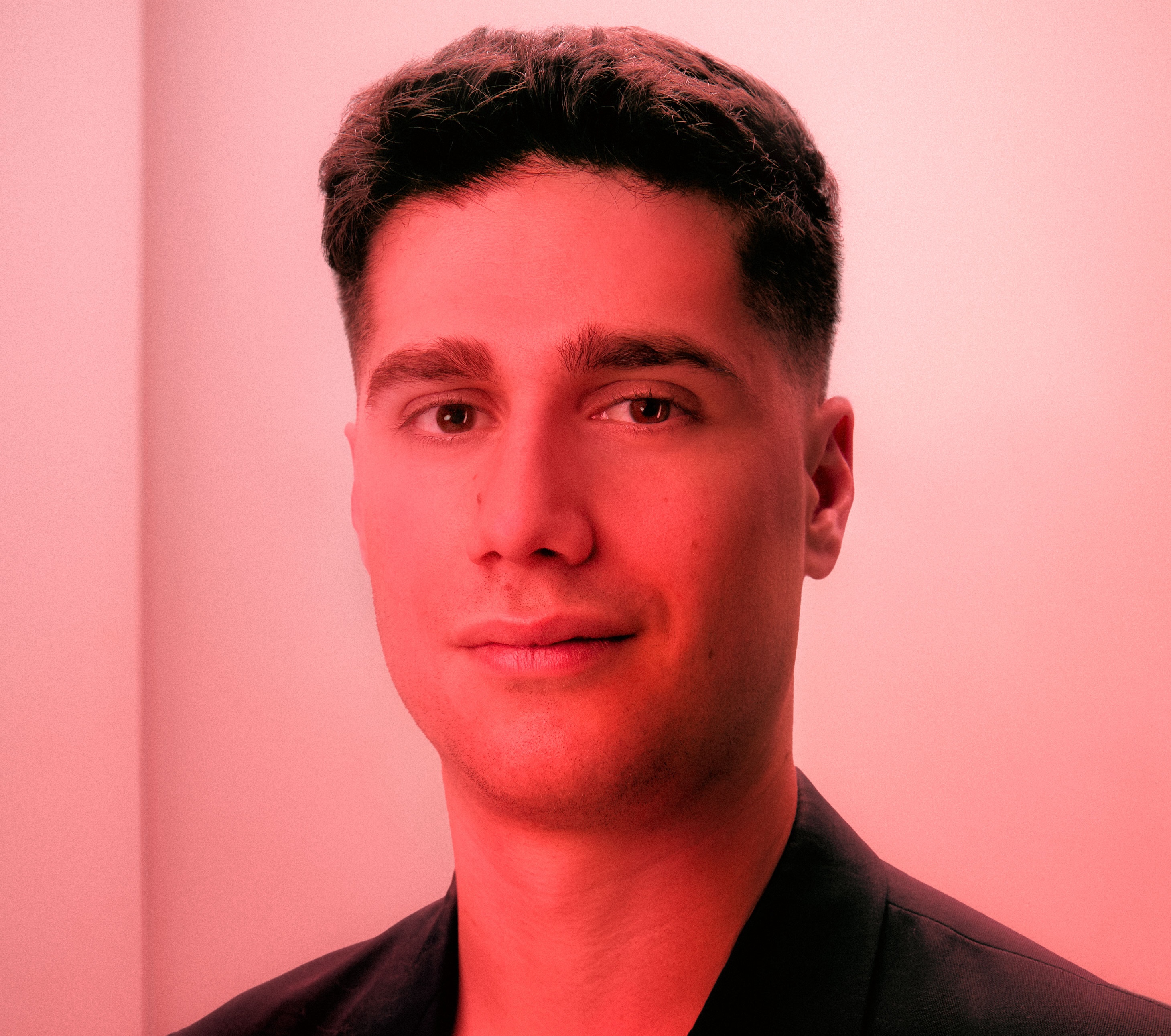 Portrait d'un homme aux cheveux courts portant un blazer noir sur fond clair avec éclairage rouge.