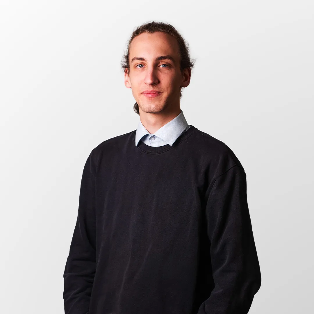 Junger Mann mit dunklem Pullover und hellblauem Hemd vor hellem Hintergrund.