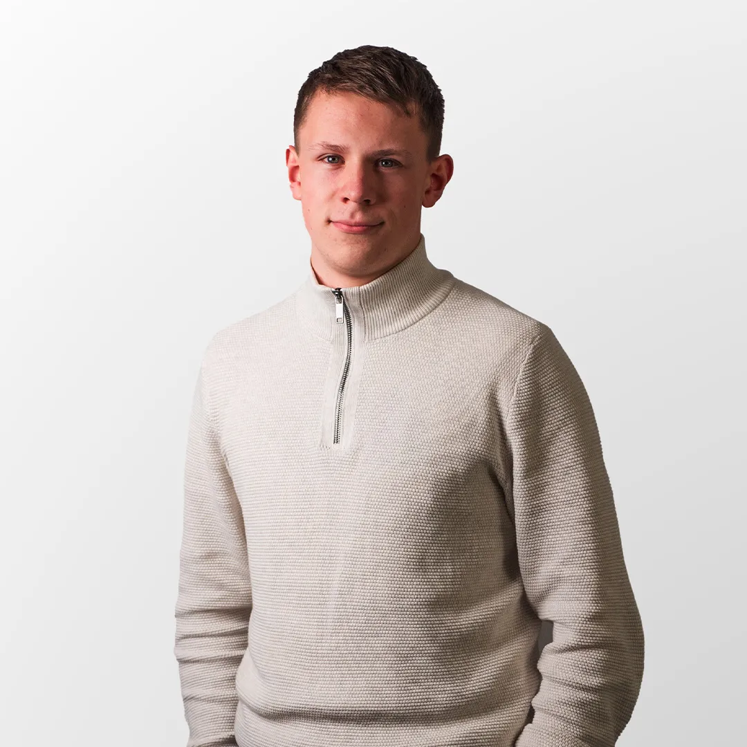 Junger Mann in einem hellen, grob gestrickten Pullover mit kurzem Reißverschluss vor einem weißen Hintergrund.