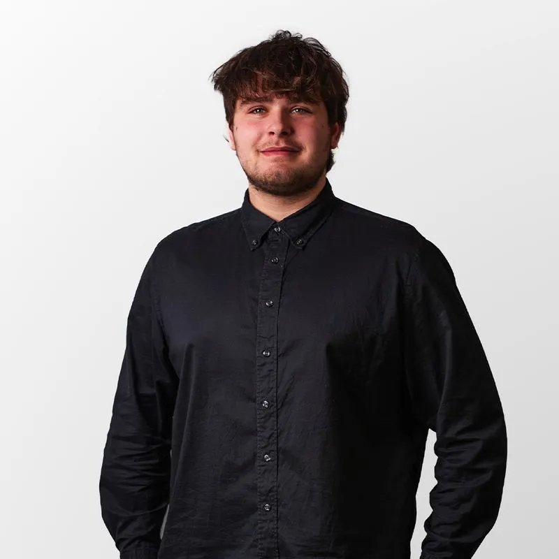 Junger Mann mit dunklem, gewelltem Haar trägt ein schwarzes Hemd und steht vor weißem Hintergrund.