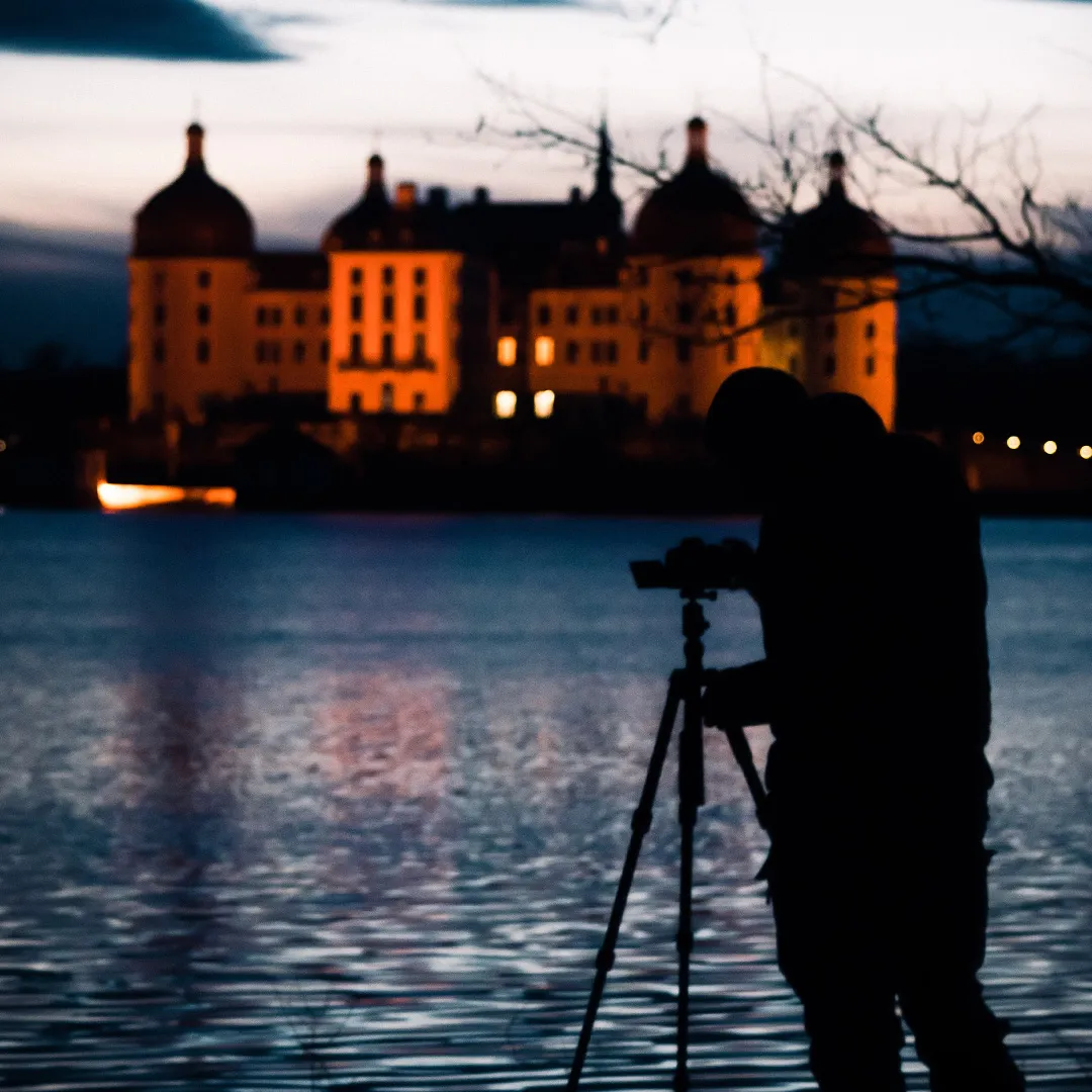 Silhouette eines Fotografen mit Stativ am Wasser bei Sonnenuntergang vor einem beleuchteten Schloss.
