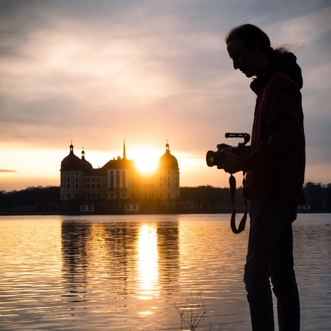 Silhouette einer Person mit Kamera vor einem See bei Sonnenuntergang, im Hintergrund ein Schloss mit reflektierendem Wasser.