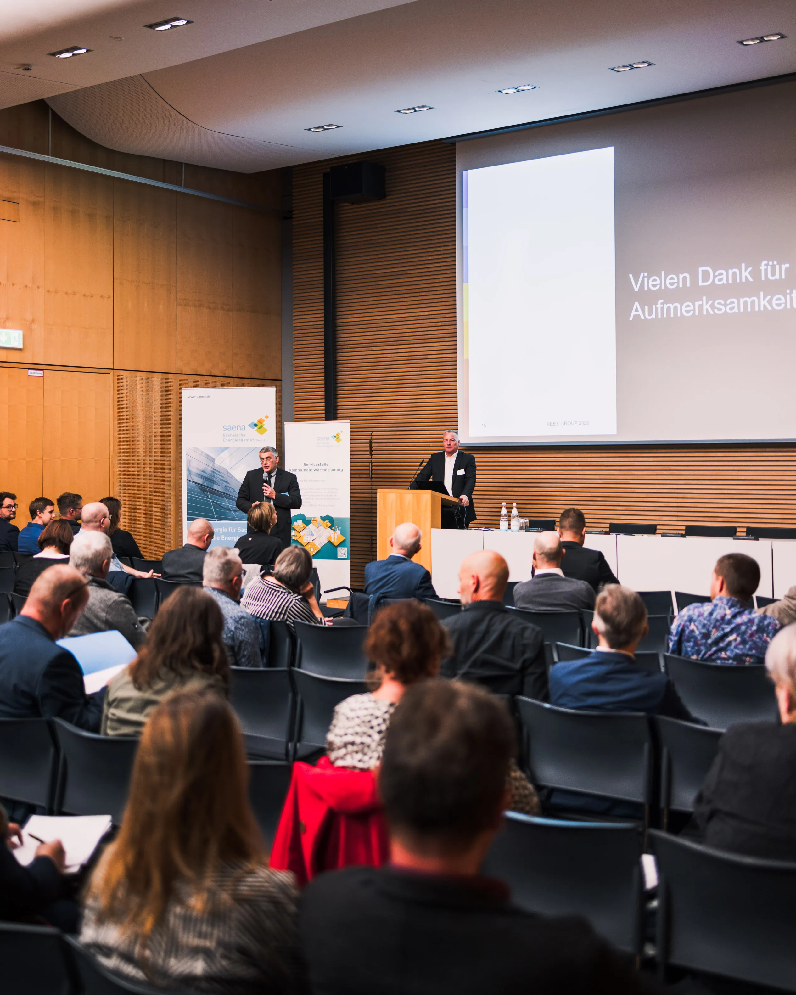 Zwei Männer sprechen auf einer Konferenz vor Publikum in einem großen Vortragssaal mit einer Projektion auf die Wand.