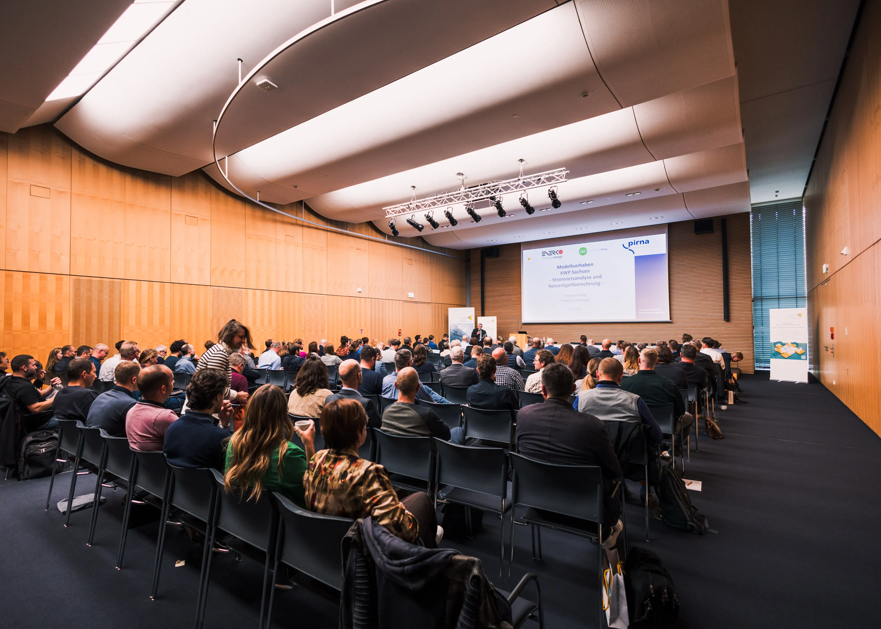 Teilnehmer einer Konferenz sitzen in einem modernen Saal und hören einem Redner vor einer Präsentation zu.