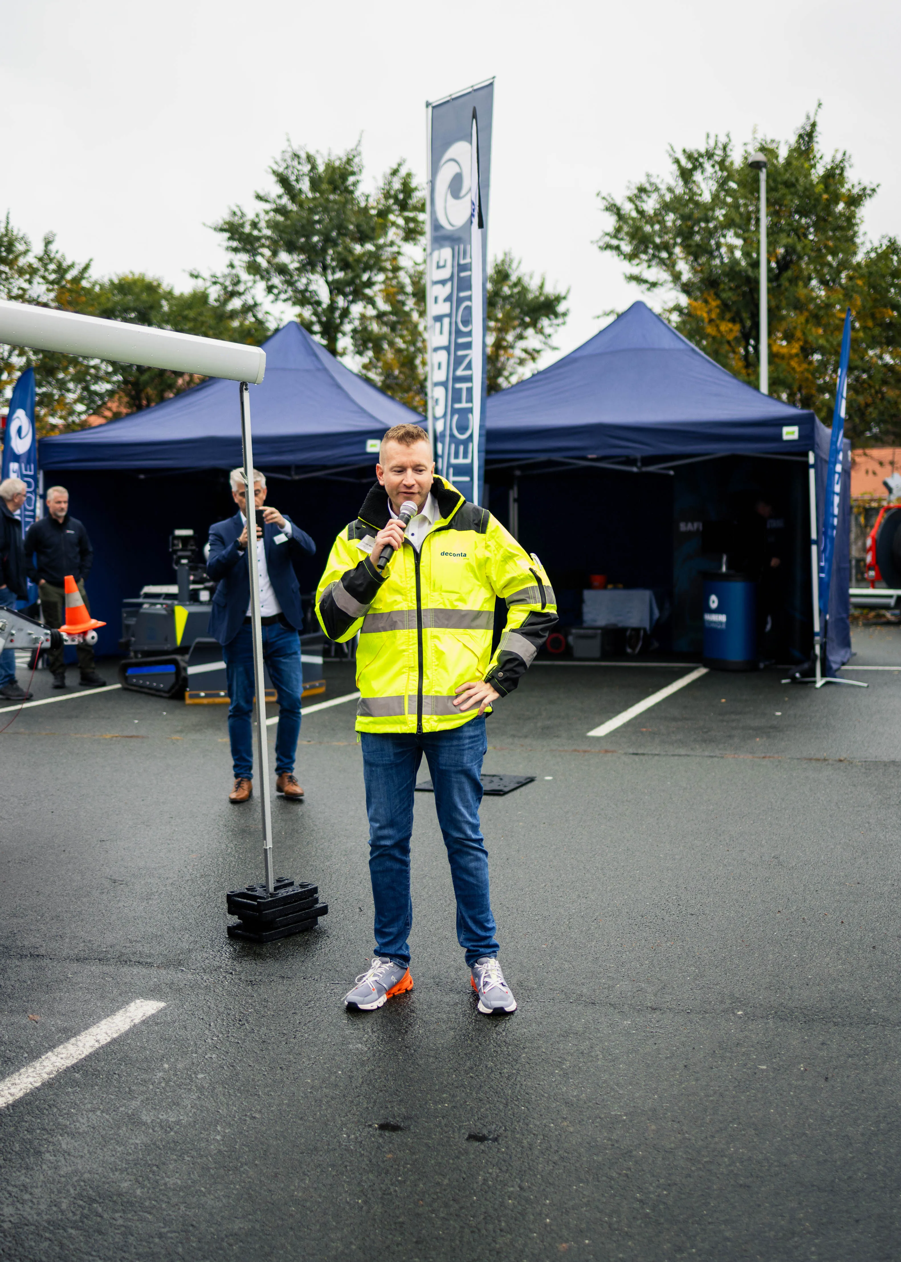Ein Mann in gelber Warnjacke hält ein Mikrofon und spricht vor einem Messestand mit blauen Zelten und Bannern im Hintergrund.