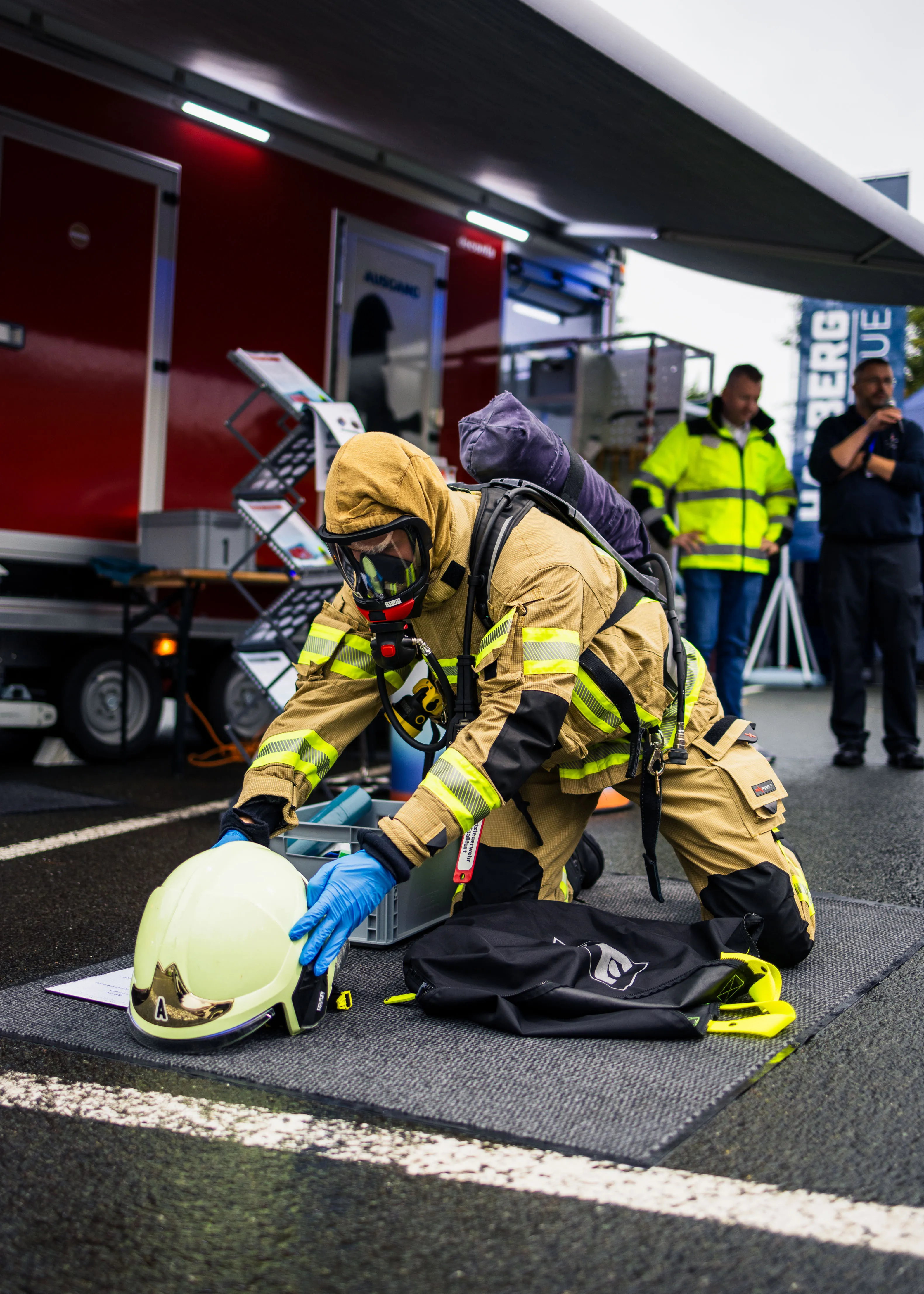 Feuerwehrmann in Schutzkleidung kniet auf Boden und greift nach einem weißen Helm vor einem roten Einsatzfahrzeug.