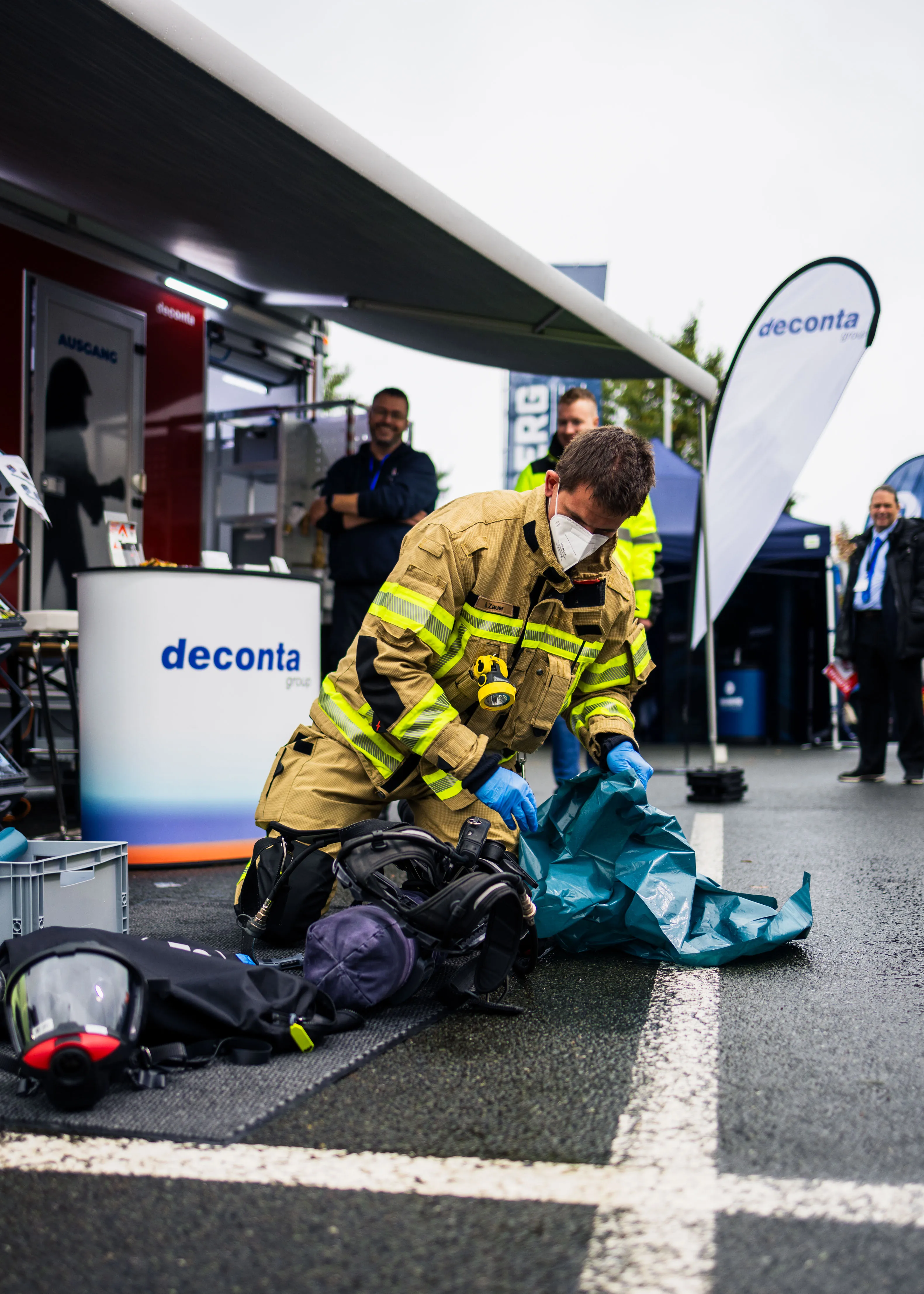 Feuerwehrmann in Schutzkleidung und Maske kniet auf Asphalt und bereitet Ausrüstung vor, im Hintergrund stehen Personen vor einem Stand mit Deconta-Logo.