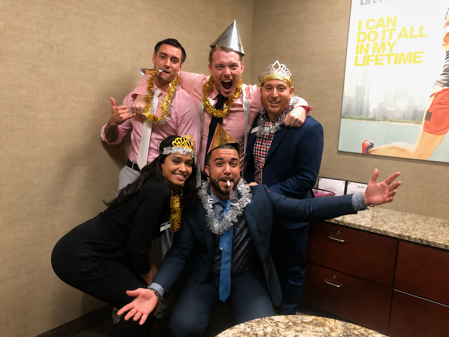 Five coworkers in party hats and tinsel smiling and posing in an office corner with a motivational poster.