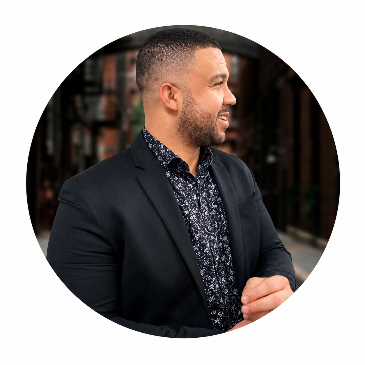 Smiling man in black blazer and floral shirt, looking right in a blurred outdoor urban setting.