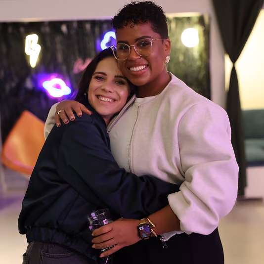 Two women smiling and hugging warmly indoors with neon lights and furniture in the background.