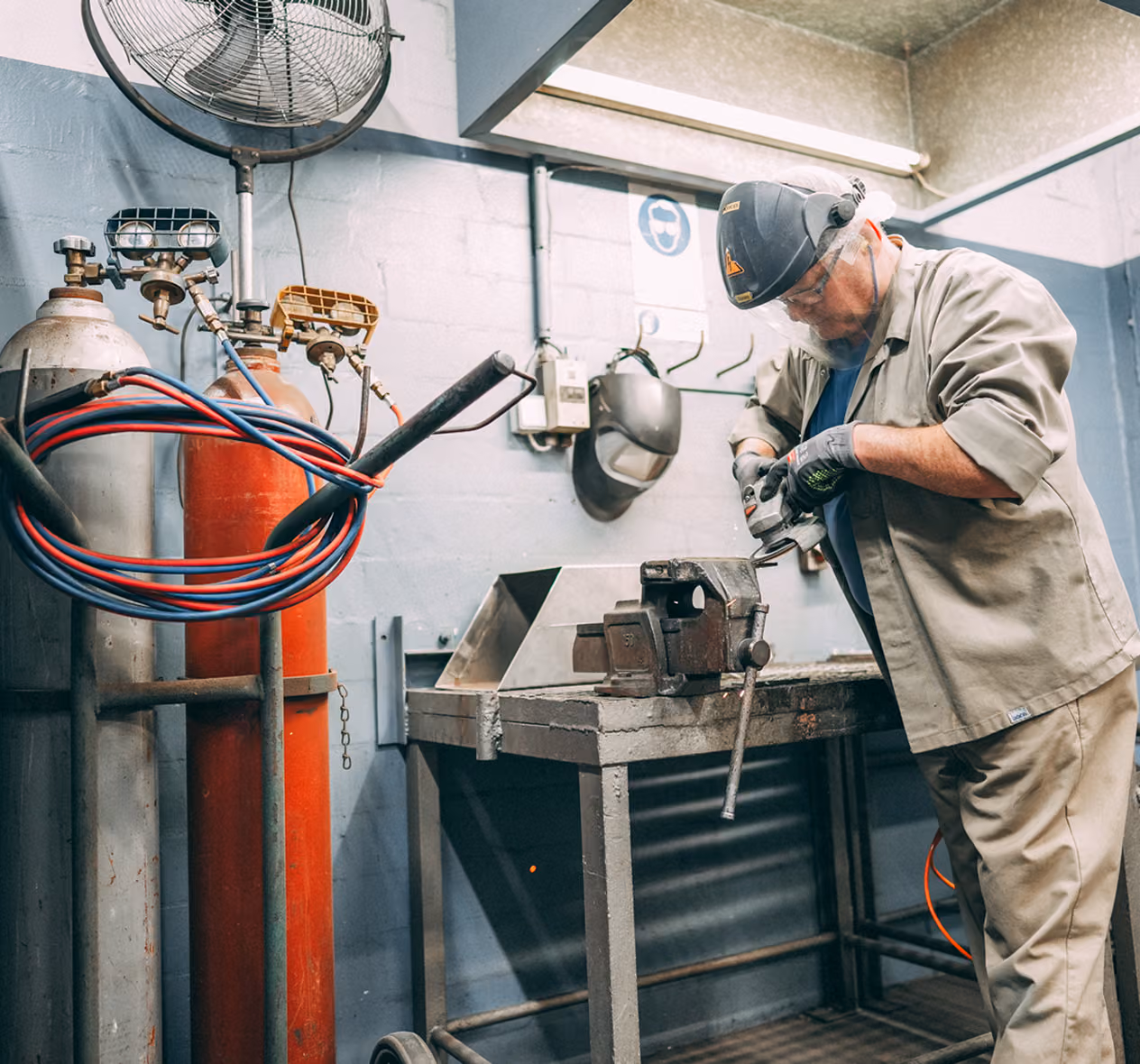 Worker wearing protective gear grinding metal clamped in a vise in an industrial workshop.