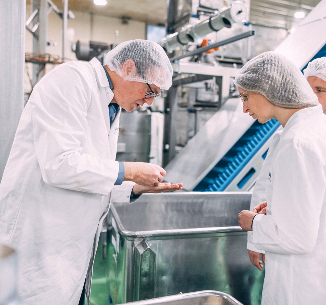 Two food factory workers in white coats and hairnets inspecting product in a metal container.