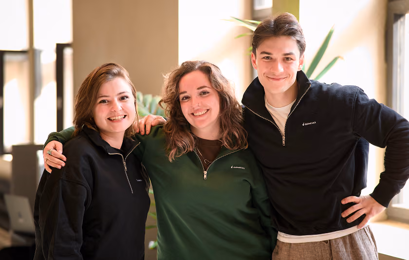 Three smiling young adults with casual black and green jackets posing indoors with arms around each other.