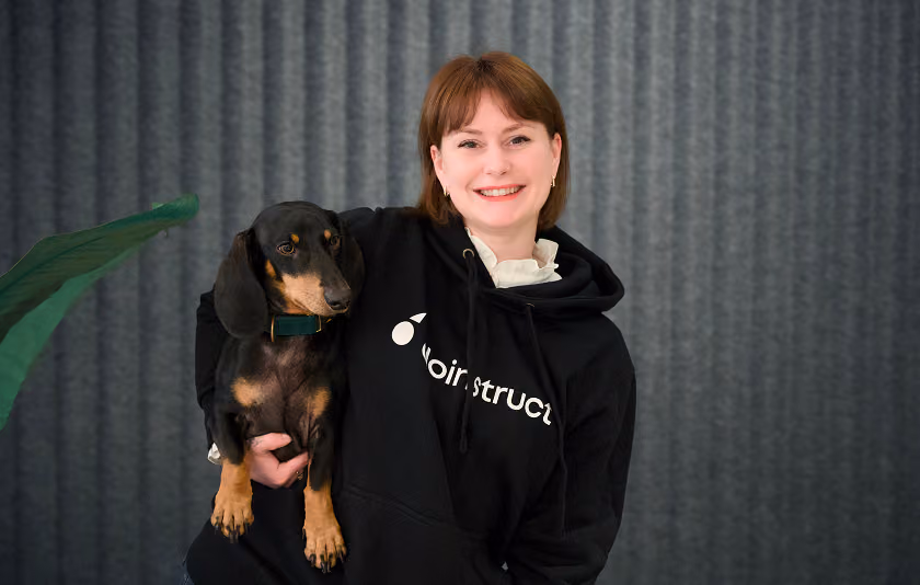 Smiling woman in black hoodie holds a black and brown dachshund against a gray curtain background.