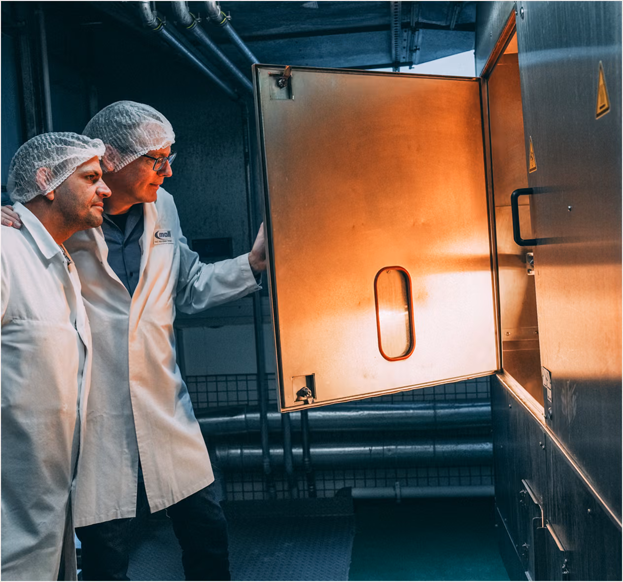 Two men in hairnets and lab coats inspecting the inside of a large industrial machine.