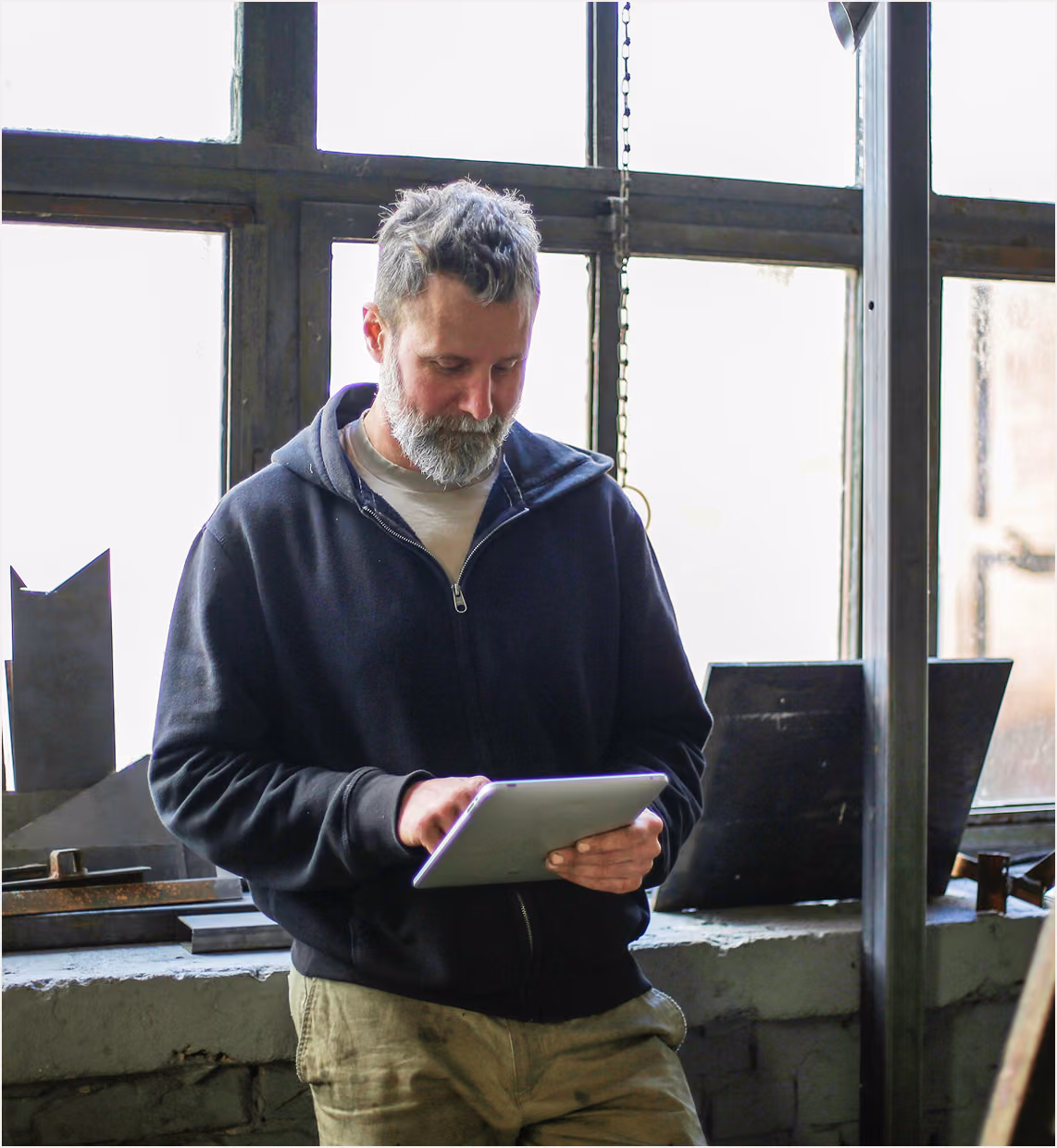 Bearded man in casual clothes using a tablet inside an industrial workshop by a window.