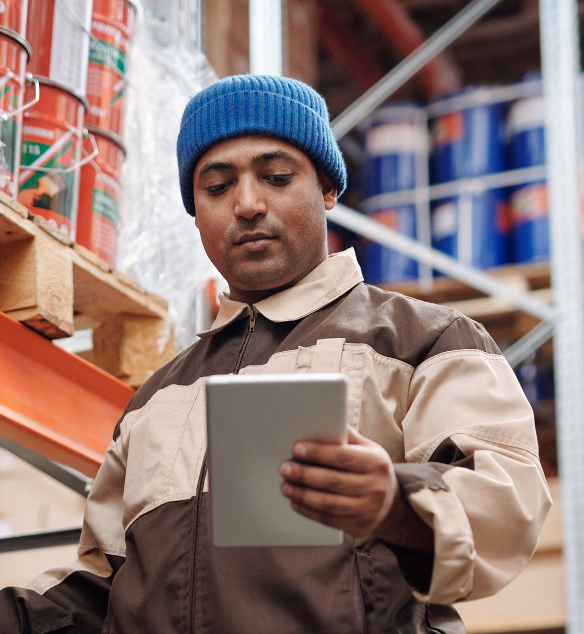 Man in work jacket and blue beanie holding a tablet in a warehouse with shelves of supplies.
