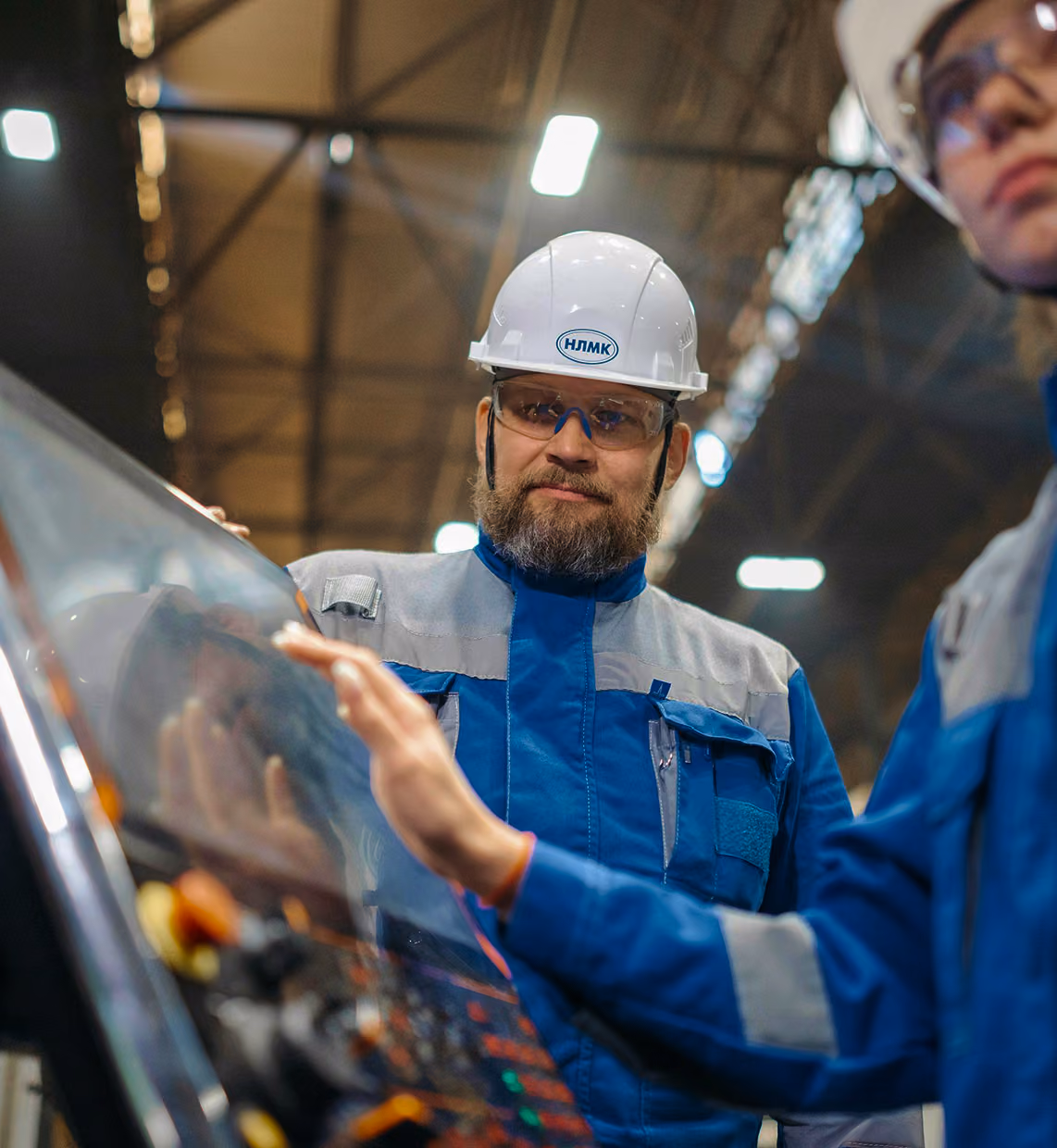 Male factory worker in safety gear operating a touchscreen control panel with female coworker nearby.
