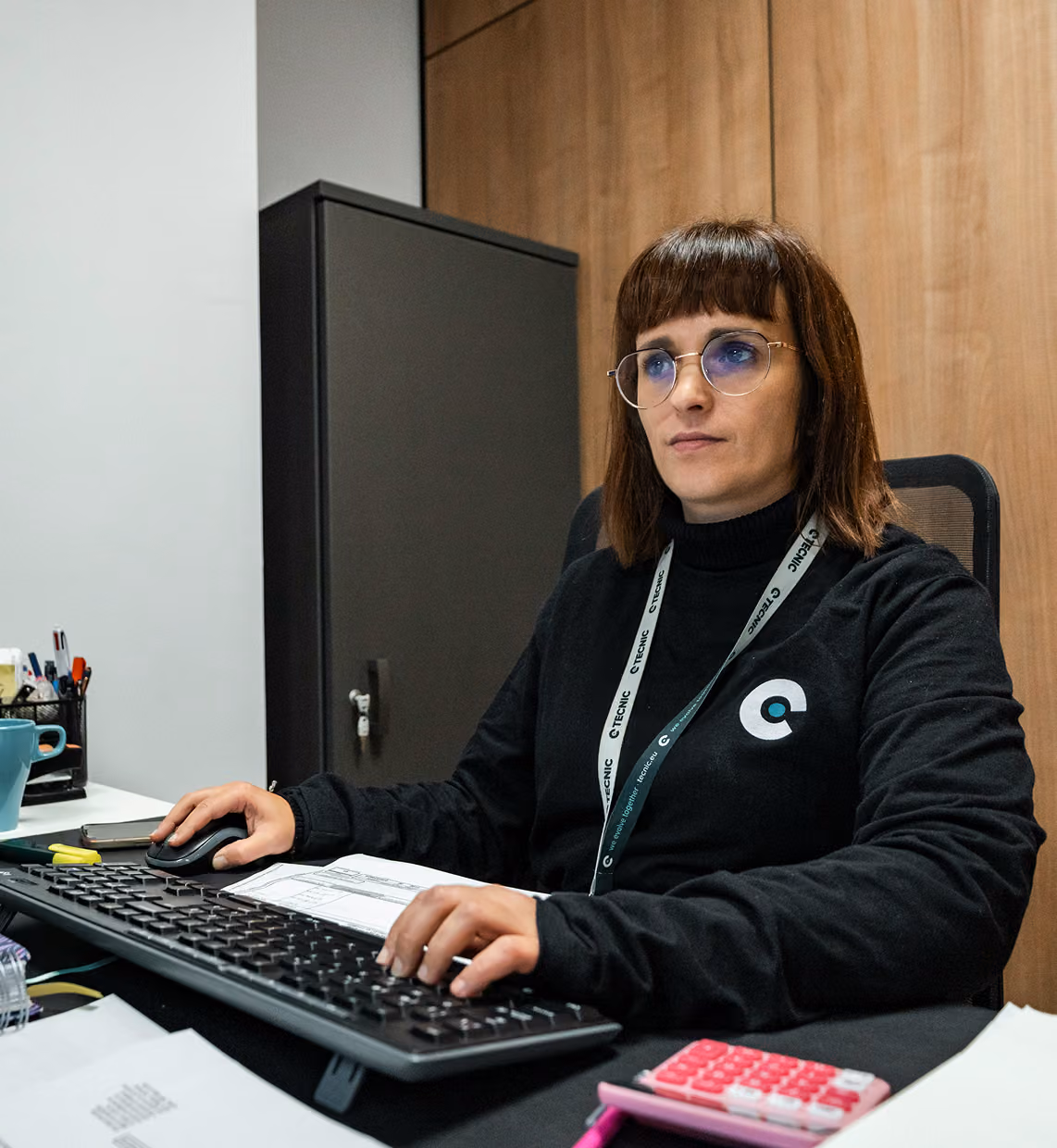 Woman with glasses seated at desk using computer keyboard and mouse in an office setting.