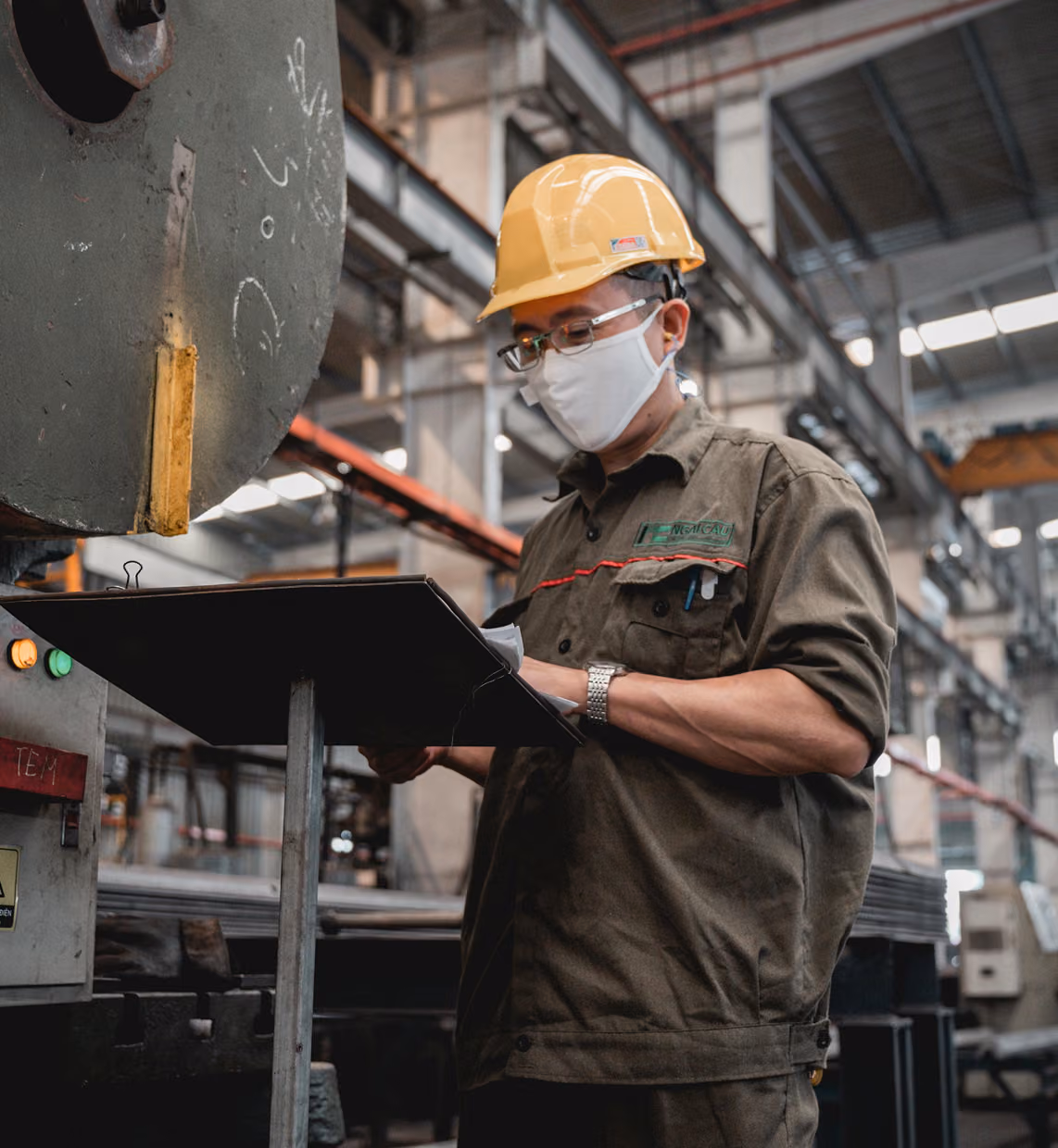 Factory worker wearing a yellow hard hat and mask, checking documents on a clipboard in an industrial setting.