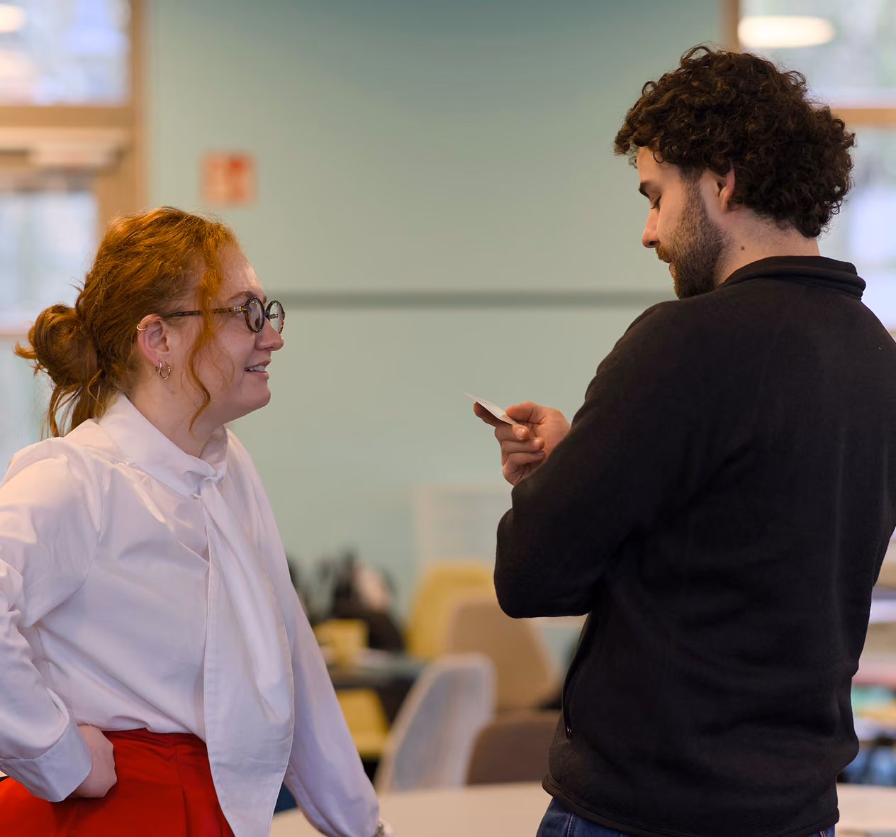 Two coworkers chatting in an office, one holding a small card while the other listens and smiles.