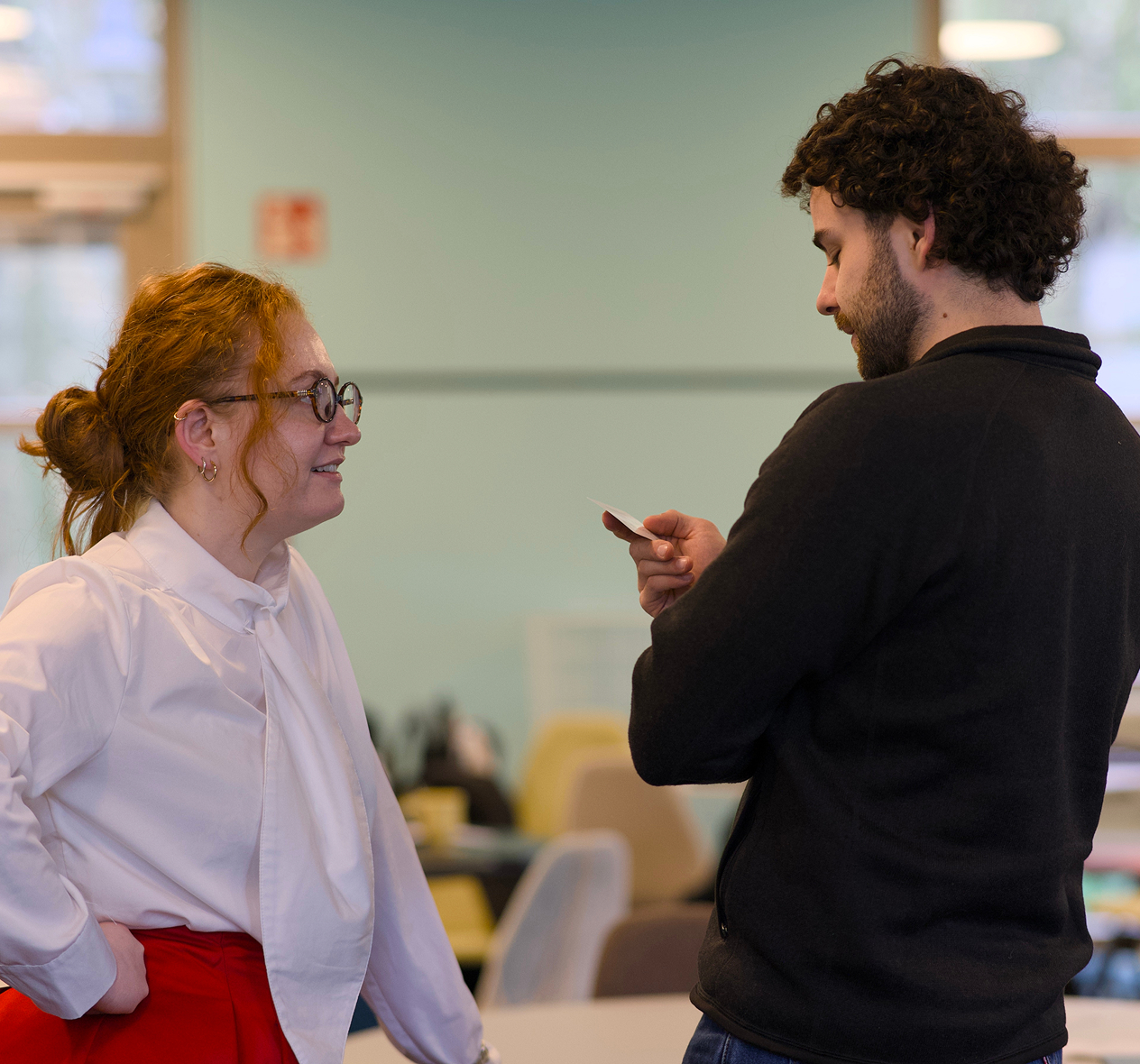 Two coworkers chatting in an office, one holding a small card while the other listens and smiles.