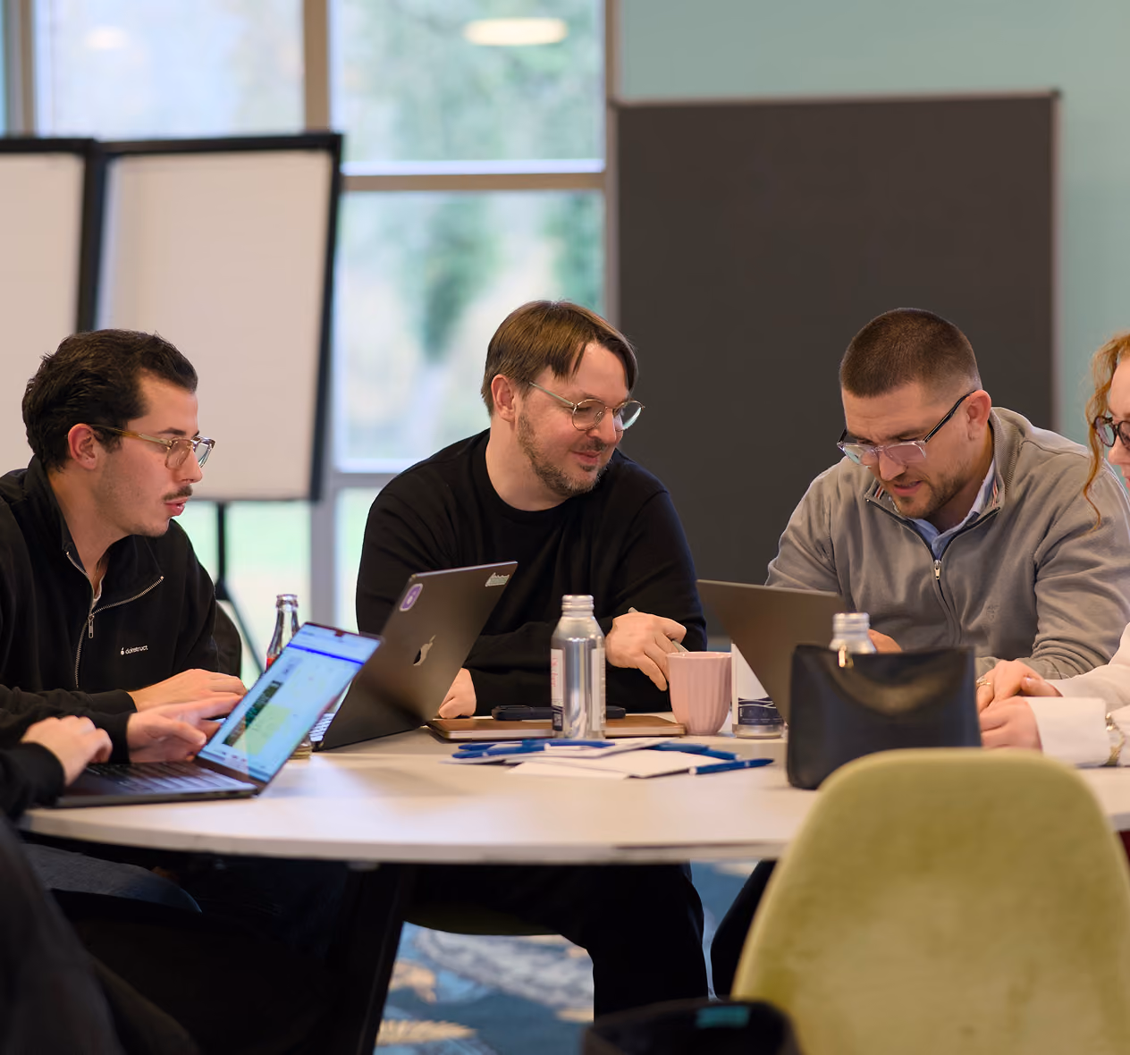Four adults with laptops and drinks collaborating around a round table in a bright office.