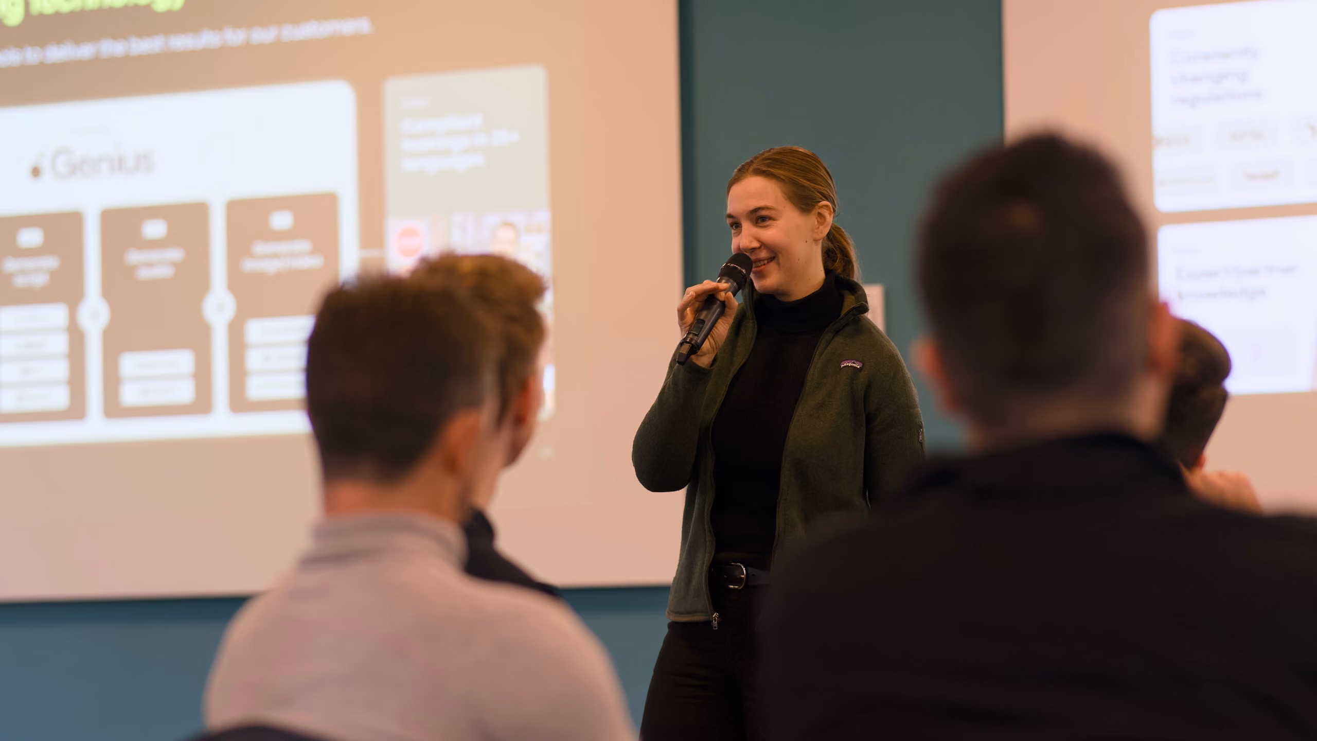 Woman speaking with microphone in front of an audience and presentation screens in a classroom or meeting room.