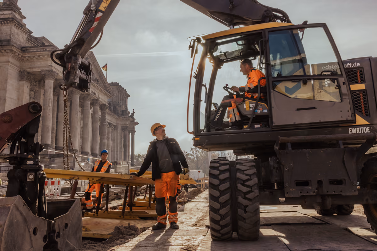 Three construction workers in safety gear at a site near a historic building with pillars.