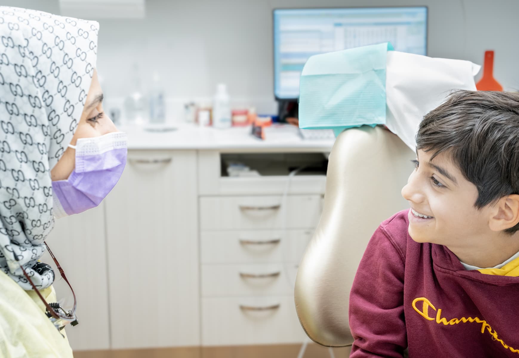 Dentist wearing a surgical cap and mask examines a young boy seated in a dental