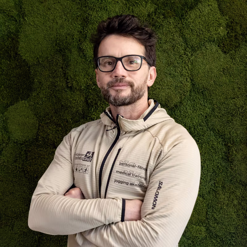Mann mit Brille und beige Jacke vor grüner Mooswand, die Arme verschränkt.