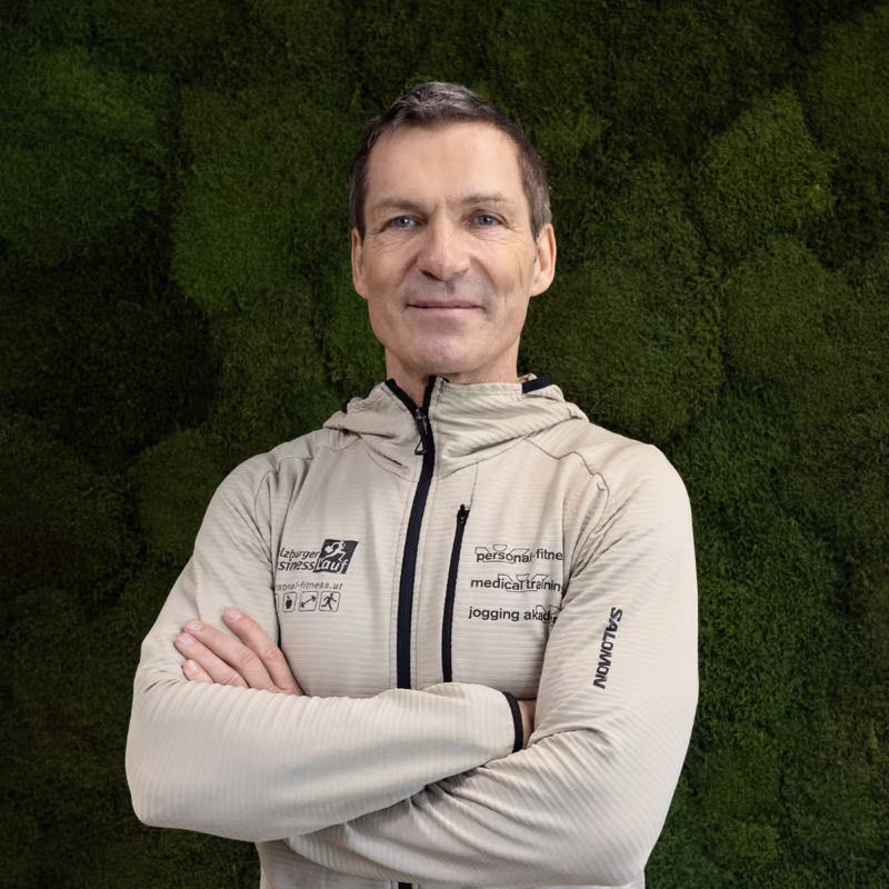 Mann vor grüner Wand in beiger Sportjacke mit verschränkten Armen lächelnd.