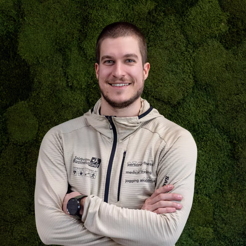Junger Mann mit beige Joggingjacke vor grünem Hintergrund, der in die Kamera lächelt.