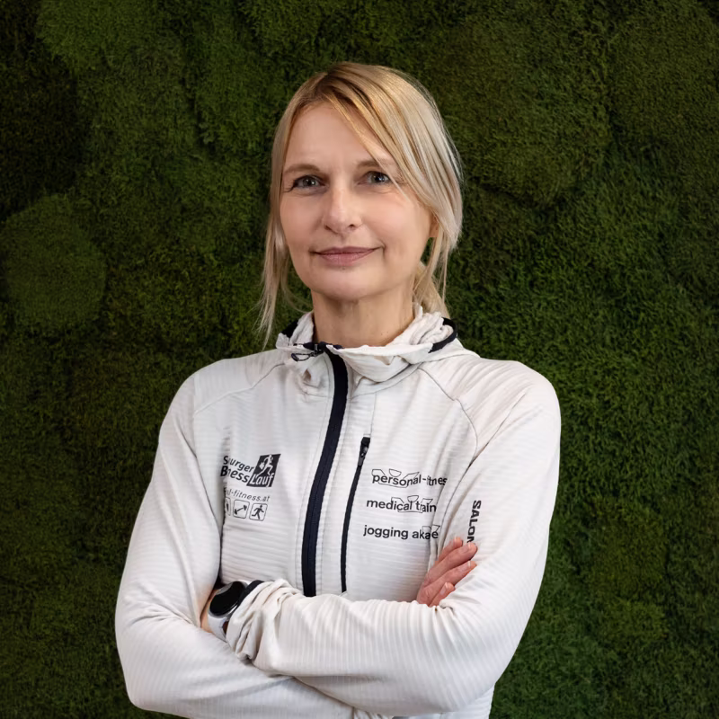 Blonde Frau in weißer Sportjacke steht vor grüner Mooswand mit verschränkten Armen.