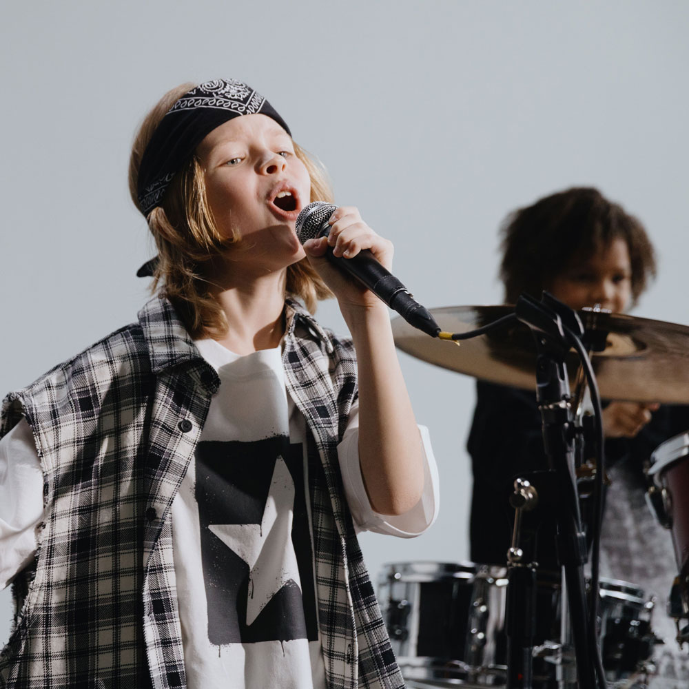 Child singing passionately into a microphone while another plays drums in the background.