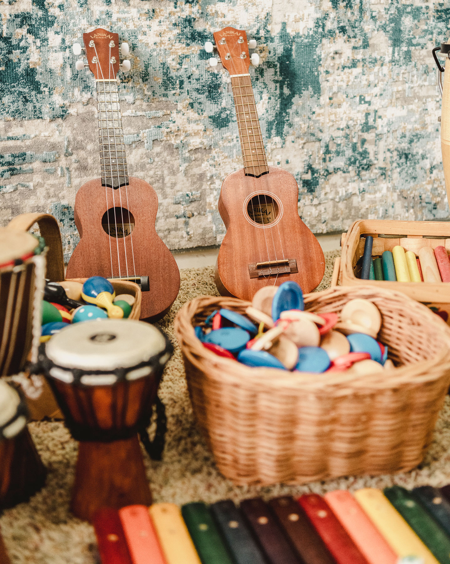 Two wooden ukuleles on a carpet next to baskets filled with colorful percussion instruments.
