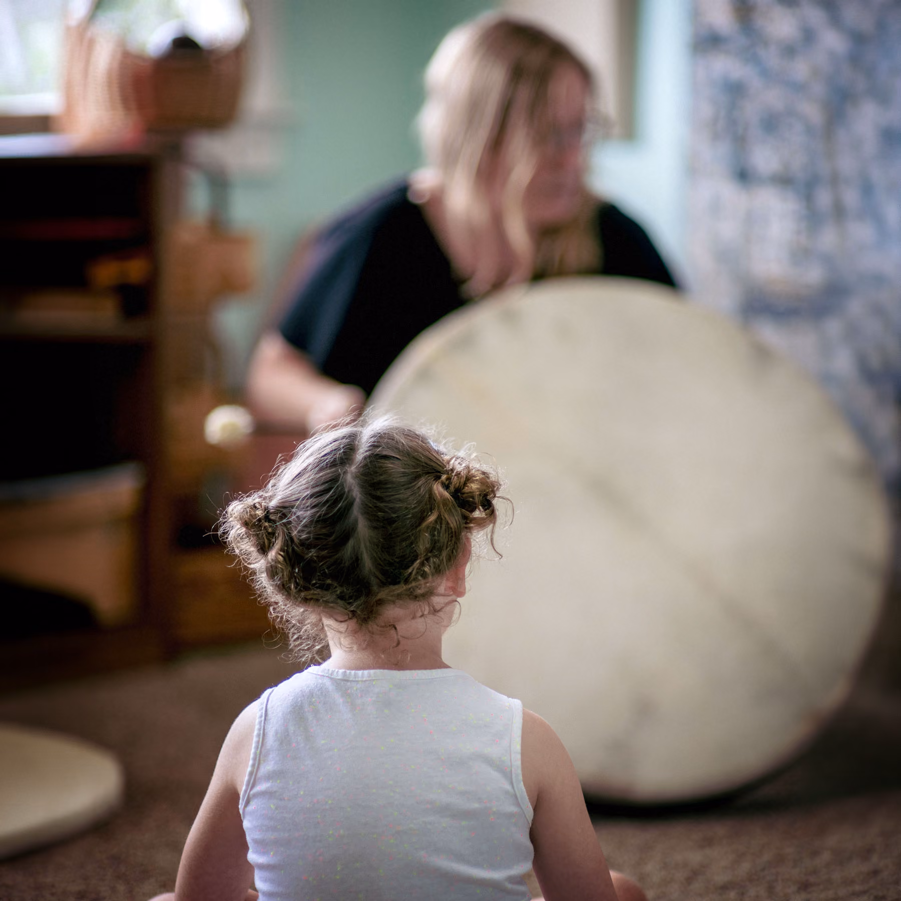 A child with curly hair in two buns sitting with their back to the camera watching an adult playing a large round drum indoors.